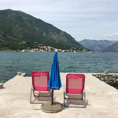 Två solstolar och ett parasoll står på en cementkaj. I bakgrunden ses hav, himmel och blånande berg. 