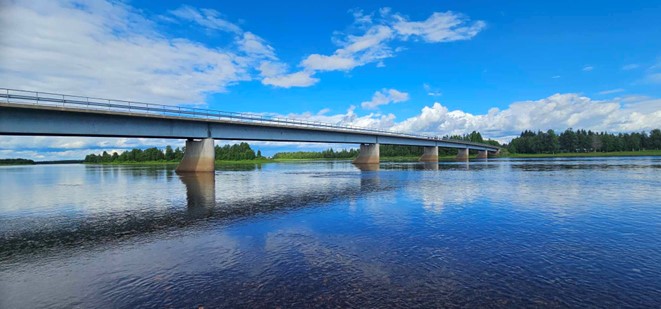 Bron vid byn Anttis, fotograferad från södra älvstranden. Bron är lång och älven bred, med skog i bakgrunden och blå himmel med lite moln. Det ser ut att vara en solig sommardag.