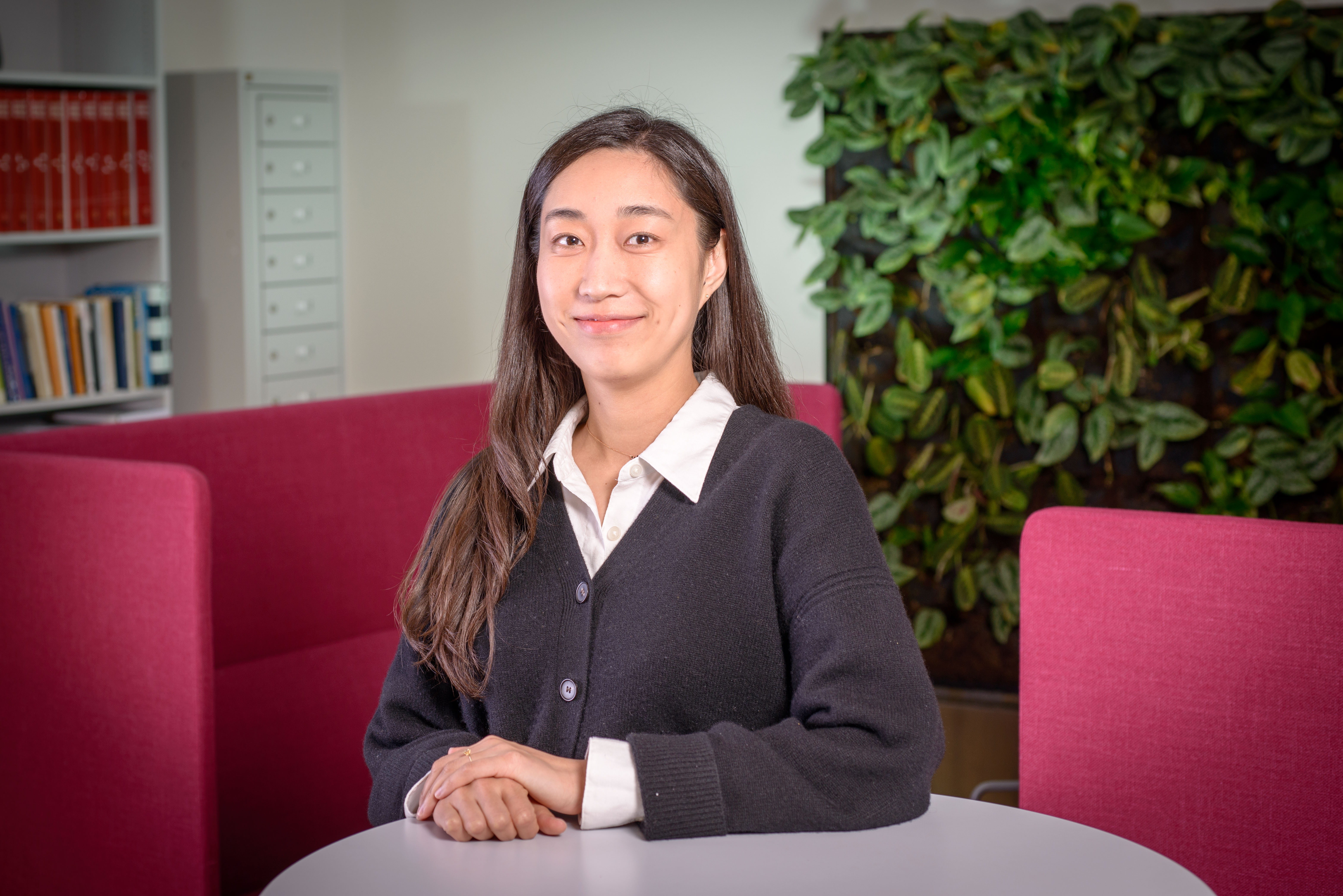 Profilbild på Hyesoo Kwon, som står vid ett runt bord med armarna vilande på bordet. Bakom finns två vinröda soffor med höga ryggstöd, en bokhylla, ett avlångt skåp och en grön växtvägg.