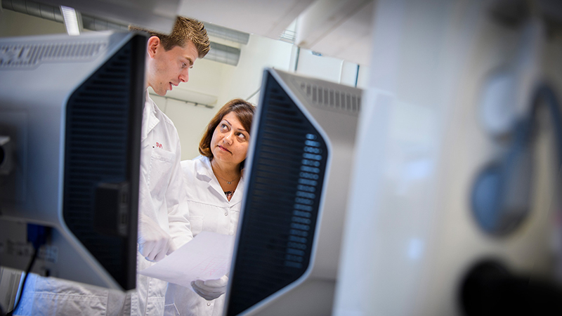 Två SVA-medarbetare i vita labbrockar ses prata med varandra bakom två bildskärmar inne i SVA:s lokaler.