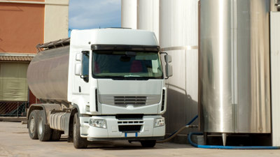 En lastbil med rostfri tank står på lantbruk vid cistern som töms på mjölk.  